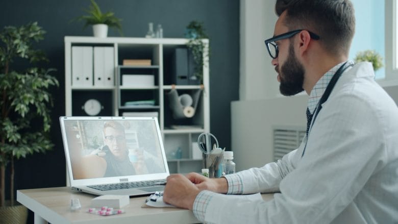 A doctor in a white coat sits at a desk, conducting a video call with a patient on a laptop in a modern office.