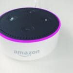 A white Amazon Echo Dot smart speaker with a black top and a glowing purple ring light, placed on a white surface.