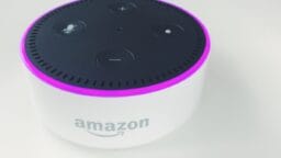 A white Amazon Echo Dot smart speaker with a black top and a glowing purple ring light, placed on a white surface.