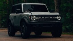 A gray Ford Bronco SUV parked on a wooded road with headlights on, surrounded by trees.