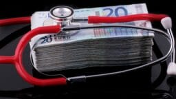 A red stethoscope rests on a large stack of 20 euro banknotes against a black background.