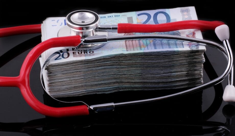 A red stethoscope rests on a large stack of 20 euro banknotes against a black background.