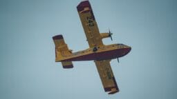 A yellow and red firefighting airplane with the number 243 is flying against a clear blue sky, viewed from below.