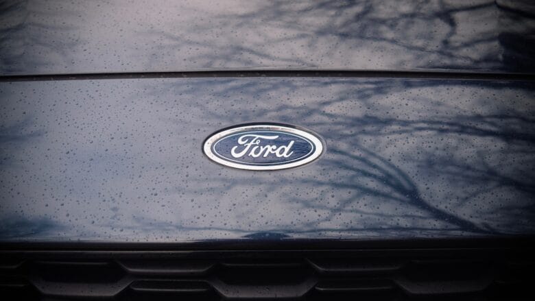 Close-up of a Ford car hood with the Ford logo in the center, water droplets, and tree reflections visible on the surface.