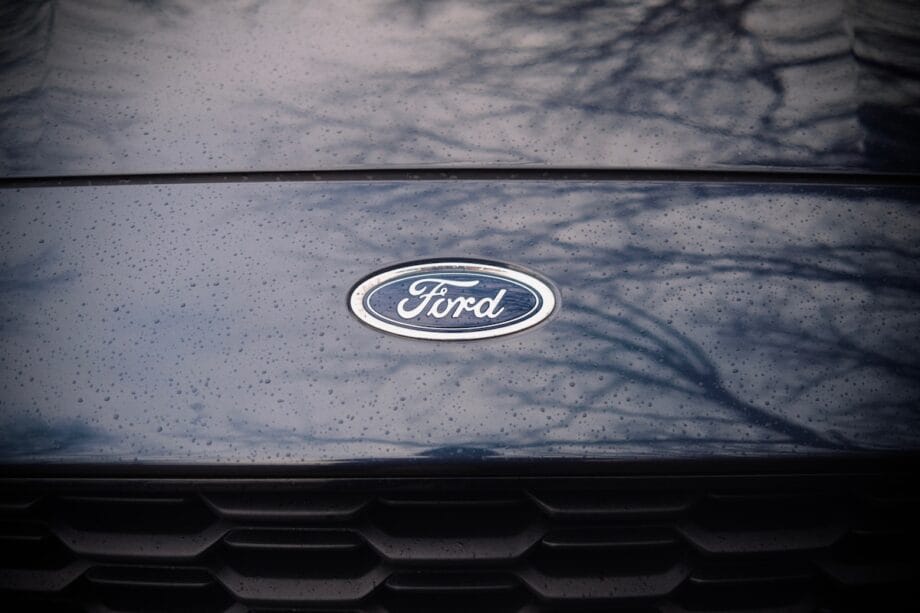 Close-up of a Ford car hood with the Ford logo in the center, water droplets, and tree reflections visible on the surface.