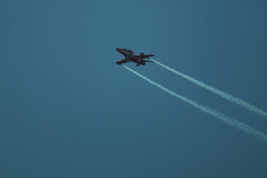 A jet aircraft with red markings flies in the sky, leaving two white vapor trails behind it.