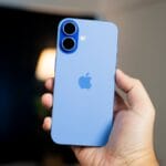 A hand holding a blue iPhone with two rear cameras, shown against a blurred indoor background.