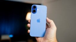 A hand holding a blue iPhone with two rear cameras, shown against a blurred indoor background.