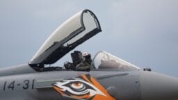 A fighter jet with cockpit open and pilot inside, featuring a tiger eye design on the nose, against a cloudy sky.
