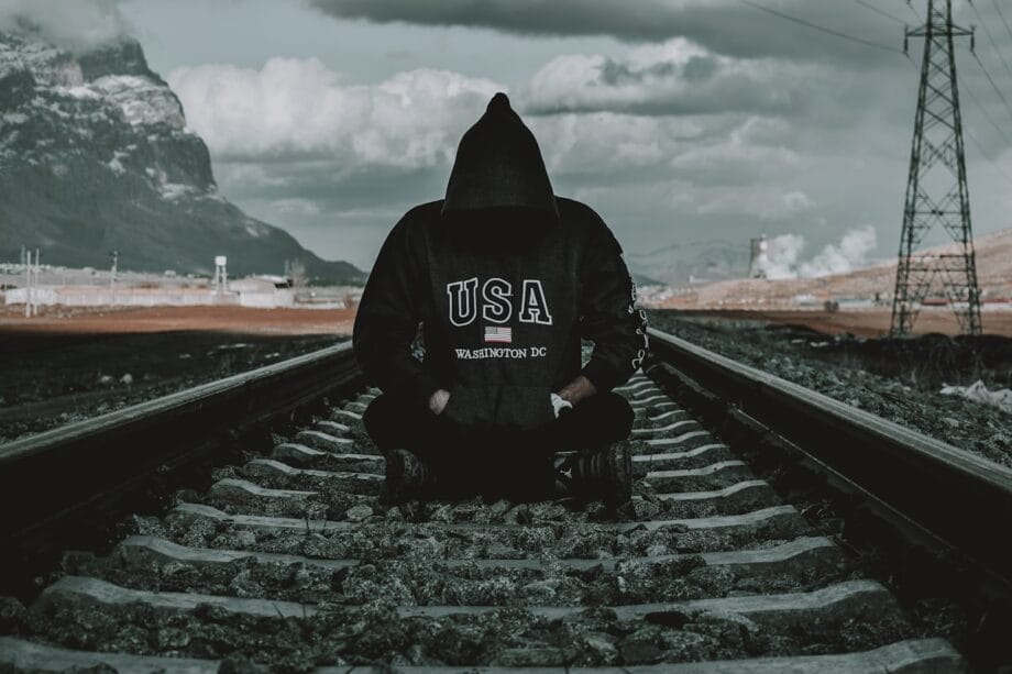 Person in a black USA Washington DC hoodie sits cross-legged on railway tracks, with mountains and power lines in the background.