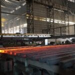 Glowing hot steel beams are processed on a conveyor inside an industrial metal factory with overhead cranes and large machinery.