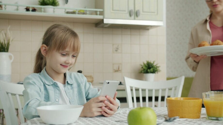 A young girl sits at a kitchen table using a smartphone while an adult brings a plate of muffins in the background.