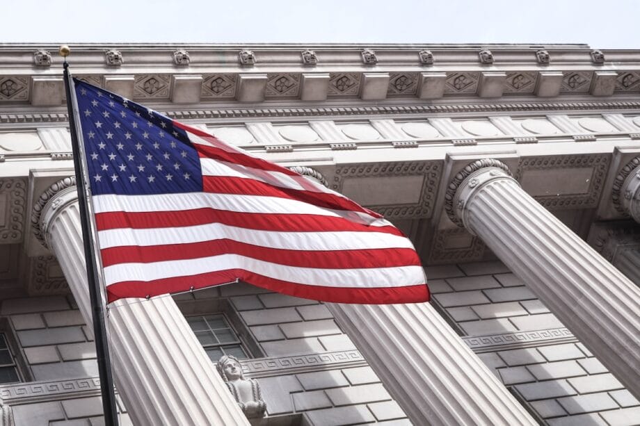 The United States flag waves in front of a large neoclassical building with tall columns.