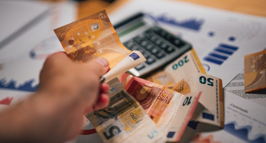 A hand counts euro banknotes near a calculator and financial charts on a desk.