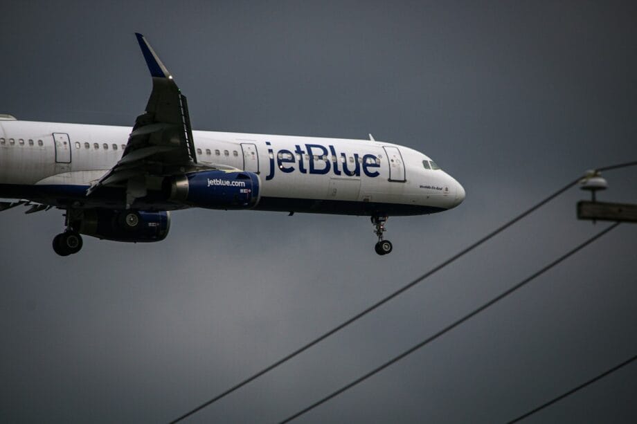 A JetBlue airplane with landing gear down flies against a dark, cloudy sky, with power lines visible in the foreground.