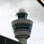Tall airport control tower with round radar dome on top, viewed from below against a cloudy sky and partial overhang in the frame.