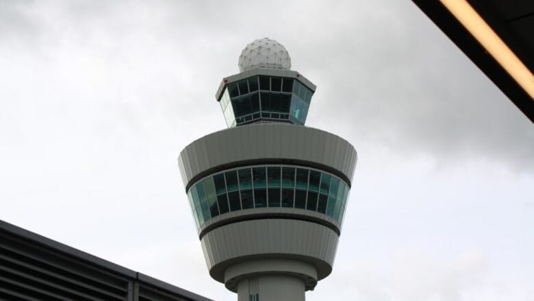 Tall airport control tower with round radar dome on top, viewed from below against a cloudy sky and partial overhang in the frame.