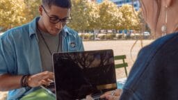 Two people sit at an outdoor table, each working on a laptop with trees and tall buildings in the background.