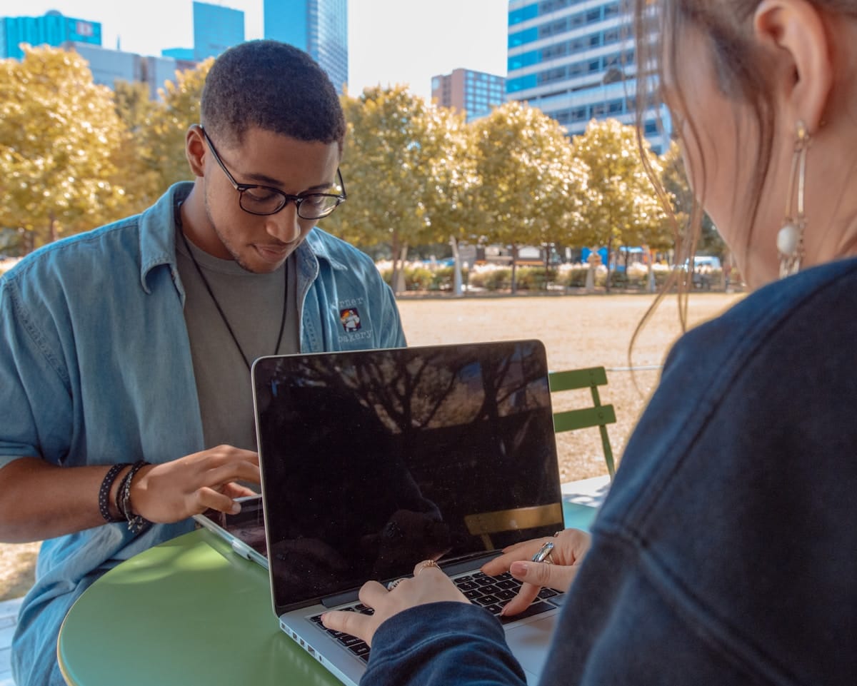 Two people sit at an outdoor table, each working on a laptop with trees and tall buildings in the background.