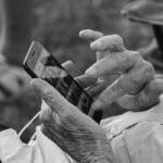 An elderly person uses a smartphone, scrolling through photos with one hand while holding the phone with the other.
