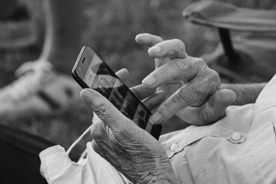An elderly person uses a smartphone, scrolling through photos with one hand while holding the phone with the other.