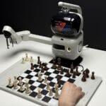 A person plays chess against a robotic arm with a display showing game information on a white table.