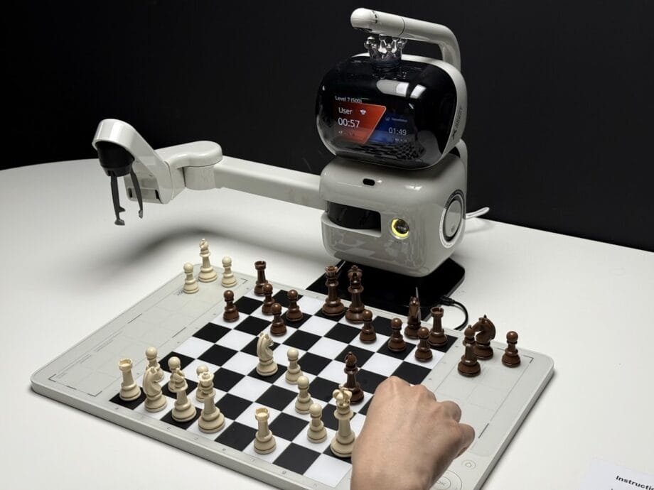 A robotic arm chess machine plays against a human hand on a chessboard, with a timer displayed on the robot’s screen.