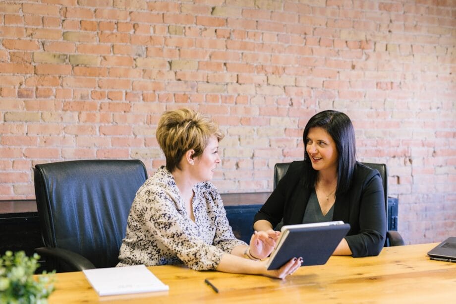 Two women sit at a conference table, discussing something on a tablet in an office with a brick wall background.