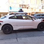A white Waymo self-driving car with sensors on the roof is parked on a city street during the day.