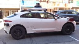 A white Waymo self-driving car with sensors on the roof is parked on a city street during the day.