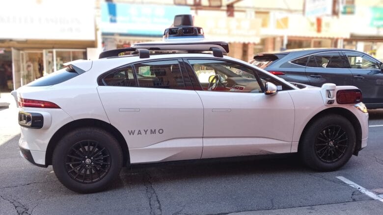 A white Waymo self-driving car with sensors on the roof is parked on a city street during the day.