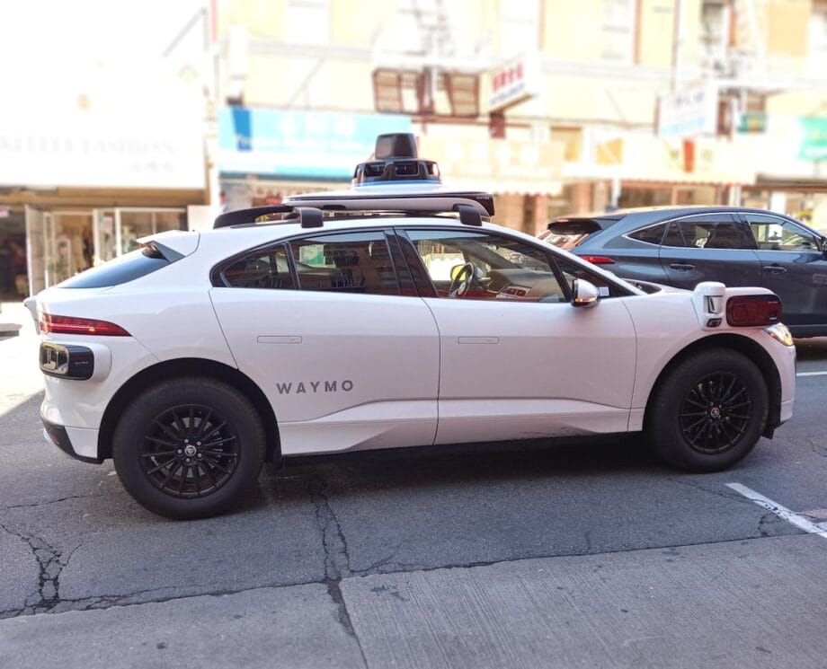 A white Waymo self-driving car with sensors on the roof is parked on a city street during the day.