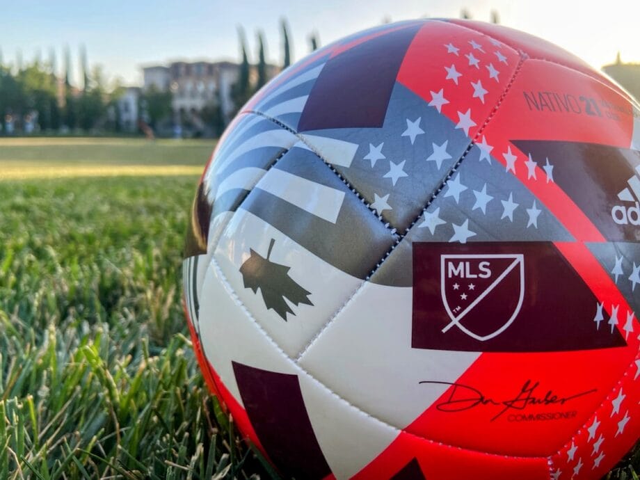 A close-up of an MLS soccer ball with a red, white, and blue design resting on a grassy field.