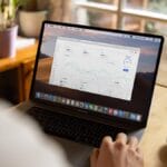 Person using a laptop displaying data analytics charts at a wooden table with a cup of coffee and a potted plant nearby.