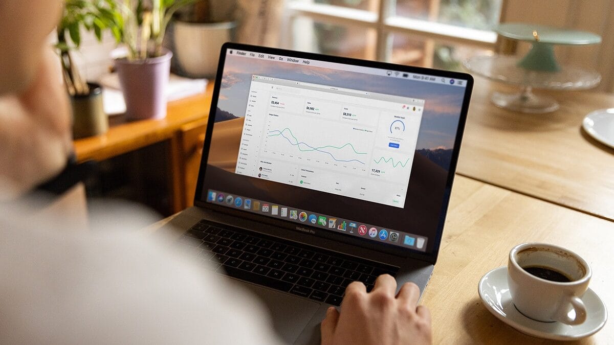 Person using a laptop displaying data analytics charts at a wooden table with a cup of coffee and a potted plant nearby.