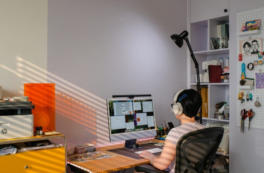 Person wearing headphones sits at a desk working on a computer in a brightly lit home office with shelves and organized supplies.