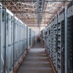 A row of industrial server racks lines a warehouse aisle, featuring exposed wiring and metal framework under a high ceiling.