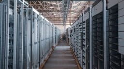 A row of industrial server racks lines a warehouse aisle, featuring exposed wiring and metal framework under a high ceiling.