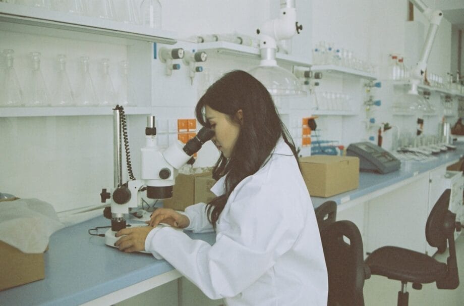 A person in a lab coat looks into a microscope at a laboratory workstation with glassware and equipment on shelves.