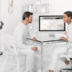 A man sits across from a woman at a desk as she shows him data on a computer screen in a bright, modern medical office.
