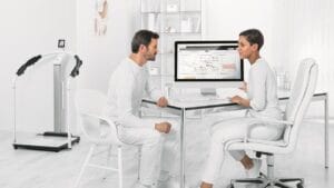 A man sits across from a woman at a desk as she shows him data on a computer screen in a bright, modern medical office.