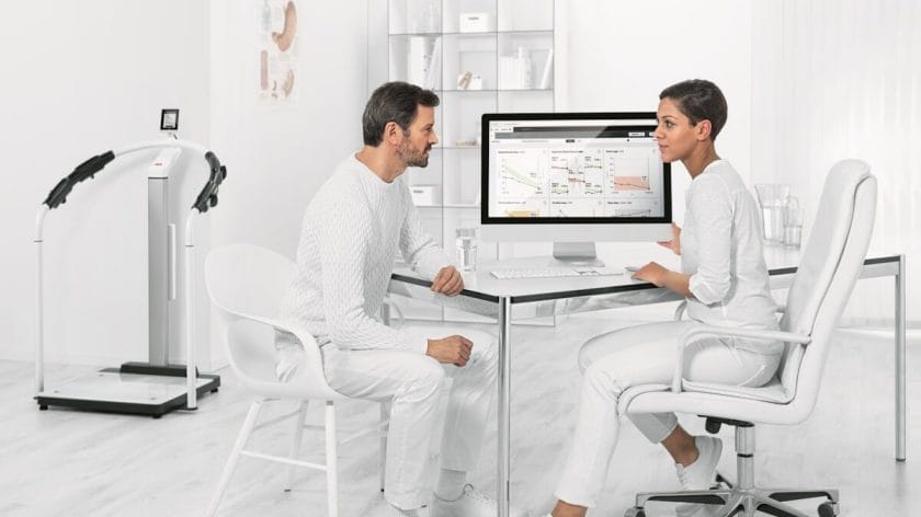 A man sits across from a woman at a desk as she shows him data on a computer screen in a bright, modern medical office.