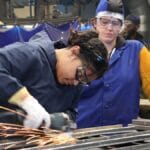 Two people in safety gear work with metal; one grinds metal, producing sparks, while the other observes.