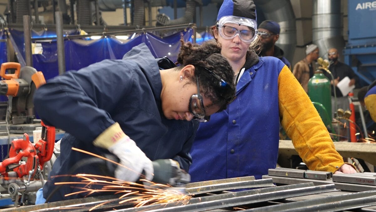 Two people in safety gear work with metal; one grinds metal, producing sparks, while the other observes.