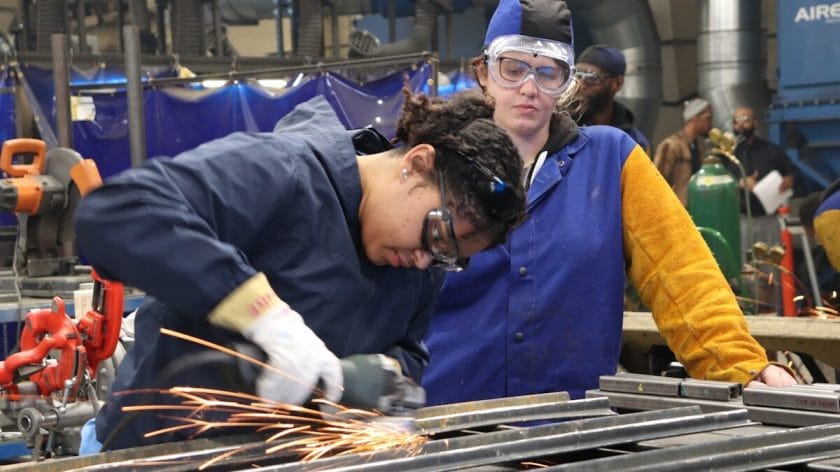 Two people in safety gear work with metal; one grinds metal, producing sparks, while the other observes.
