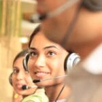 Two women wearing headsets work in a call center, with another person in the foreground out of focus.