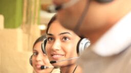Two women wearing headsets work in a call center, with another person in the foreground out of focus.
