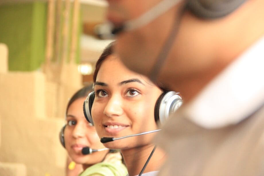 Two women wearing headsets work in a call center, with another person in the foreground out of focus.
