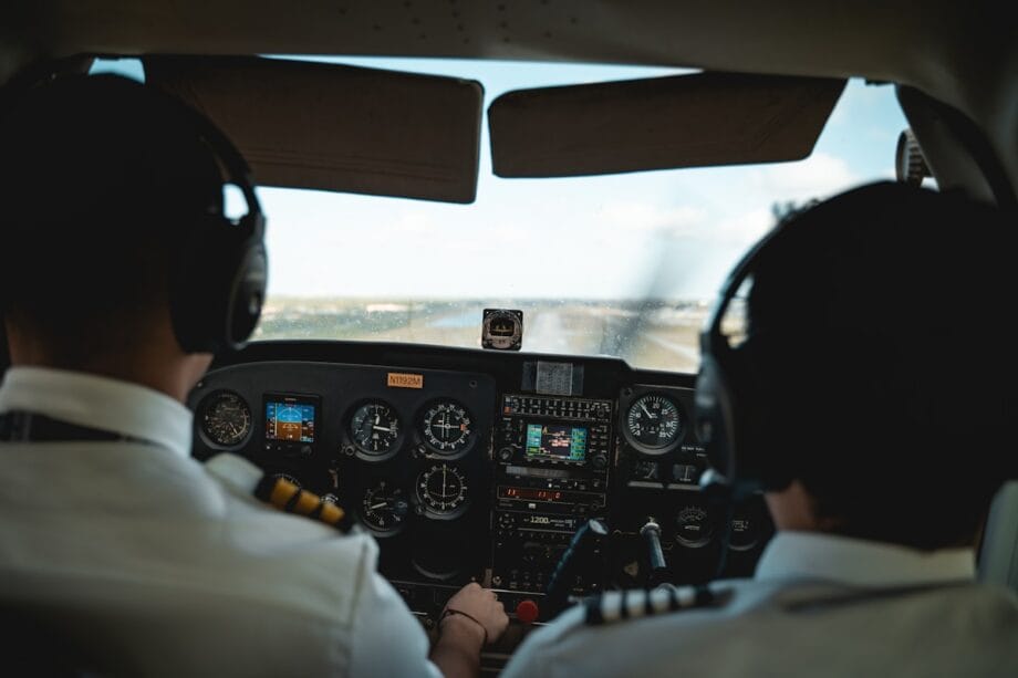 Two pilots wearing headsets sit in a cockpit, preparing to land a small aircraft, with runway visible through the windshield.