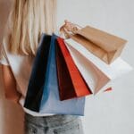 A person with long blonde hair holds several colorful paper shopping bags over their shoulder against a plain background.